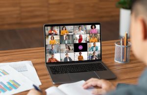 A man sits at a desk in a home office and uses a laptop to participate in a video conference meeting. The laptop screen displays a grid of video windows featuring a diverse group of people